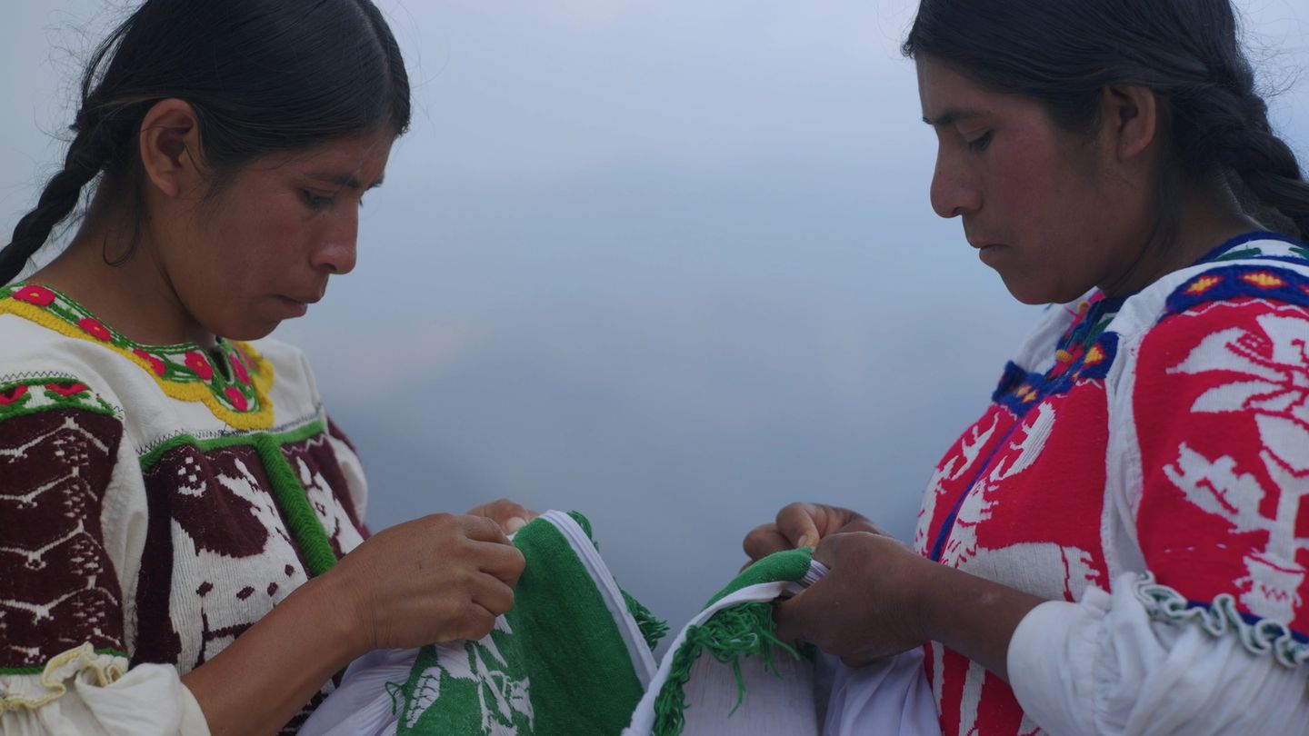 Dos mujeres bordan juntas al aire libre, con la niebla al fondo.