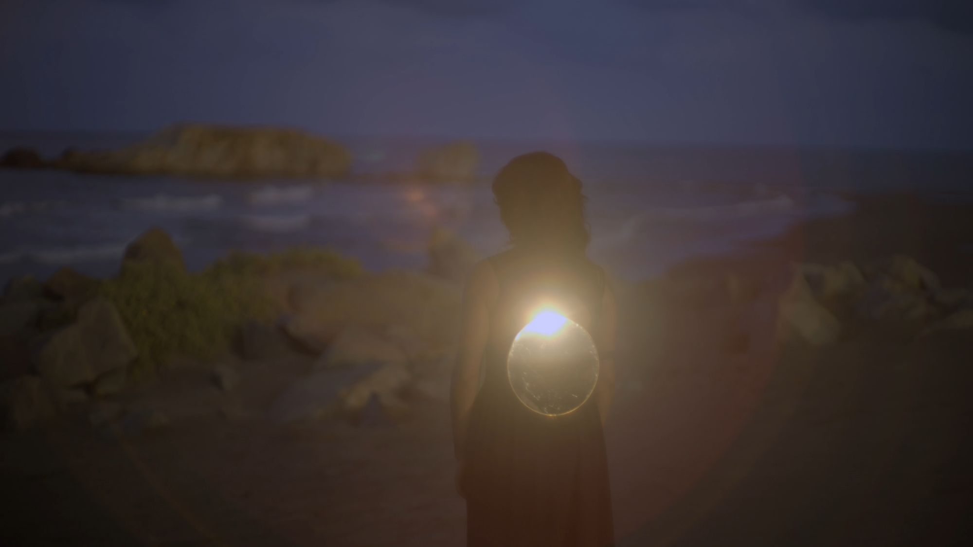 Una mujer sostiene una fuente de luz frente al mar; el sol se filtra entre sus manos.