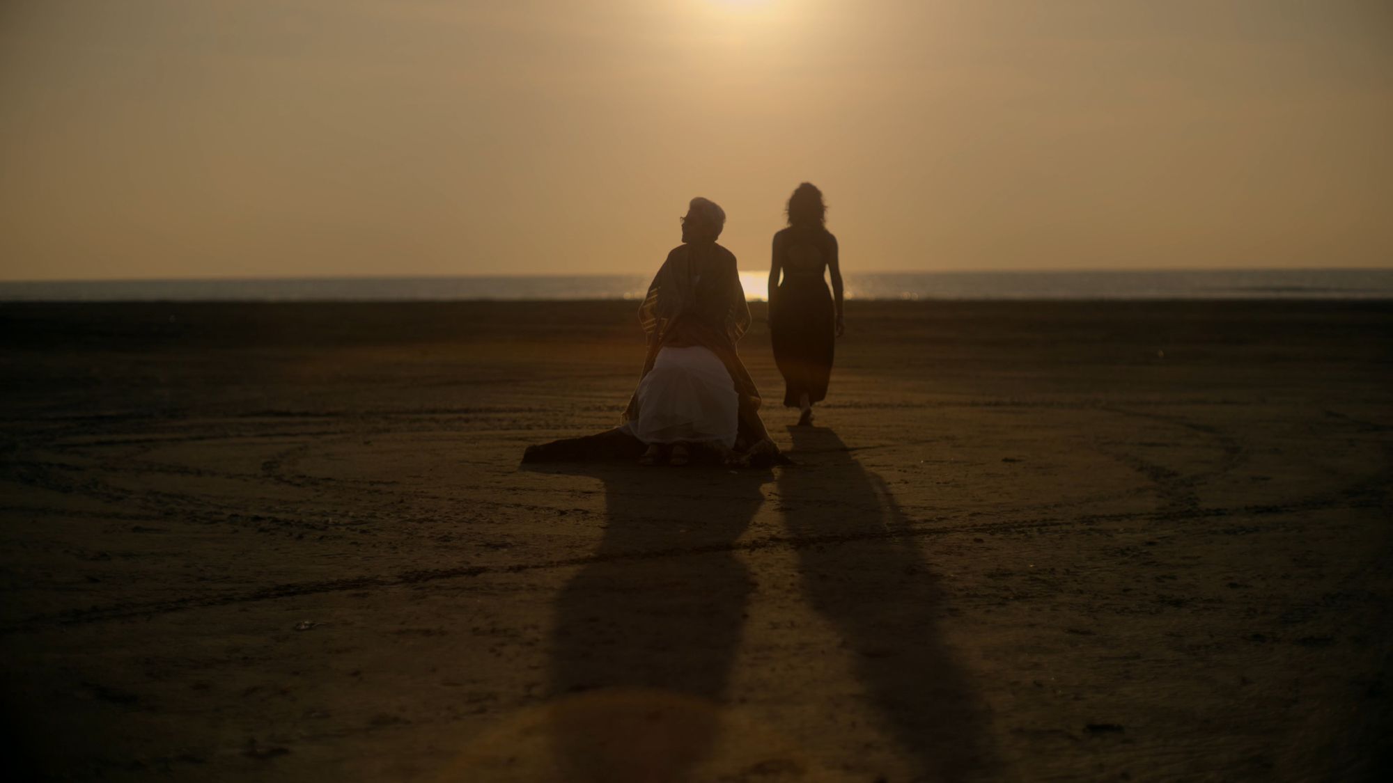 Dos figuras a contraluz del sol bajo; sombras largas sobre la arena mojada.